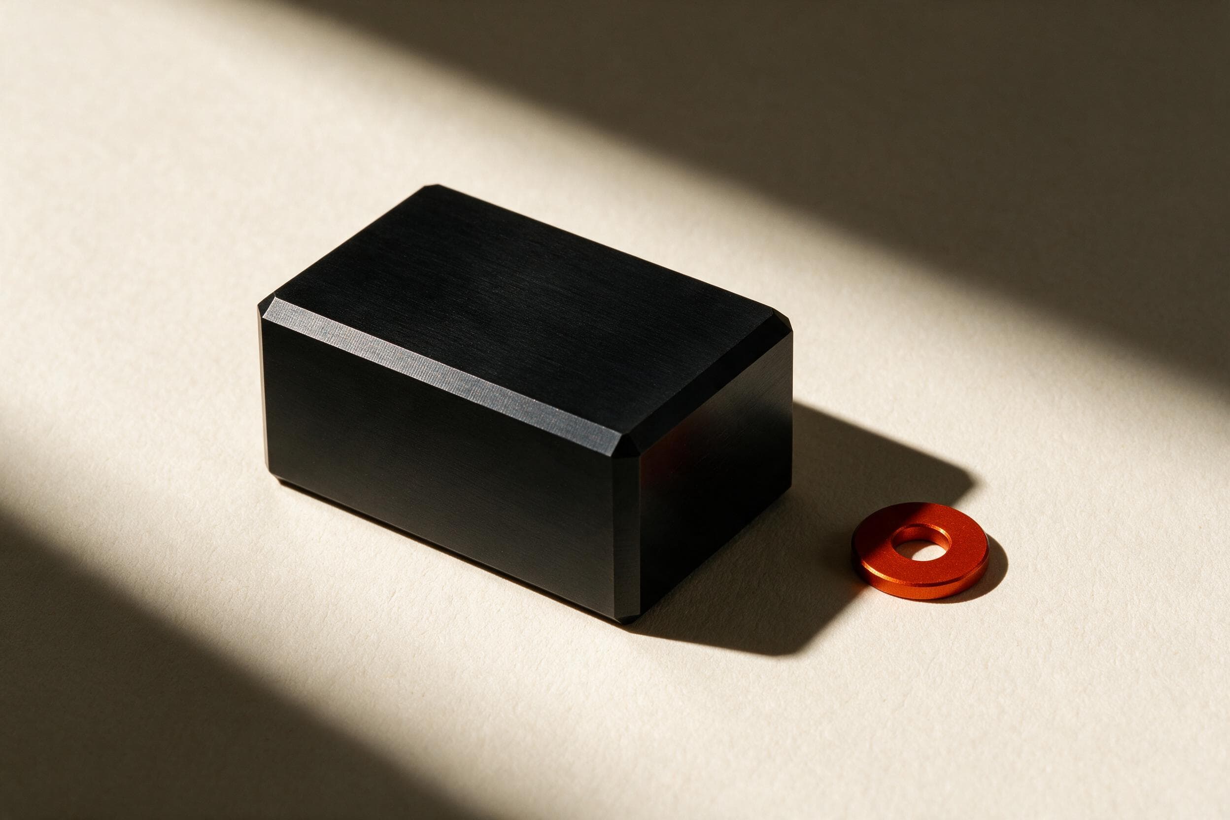A machined matte-black aluminium block with a chamfered edge catching hard light, beside a single orange anodised washer.