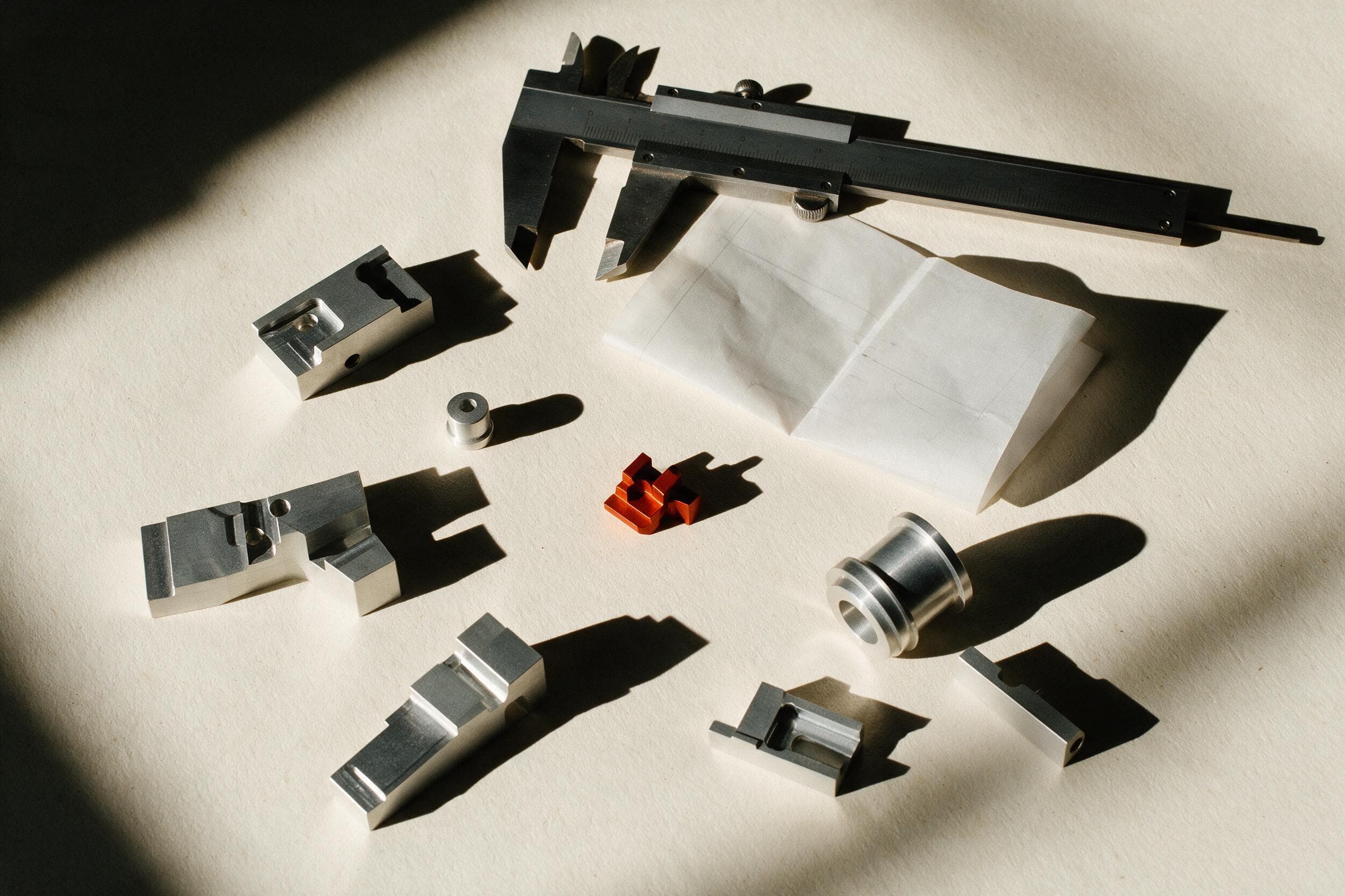 Machined aluminium components, a steel caliper and one orange anodised part laid out on a warm off-white workbench.